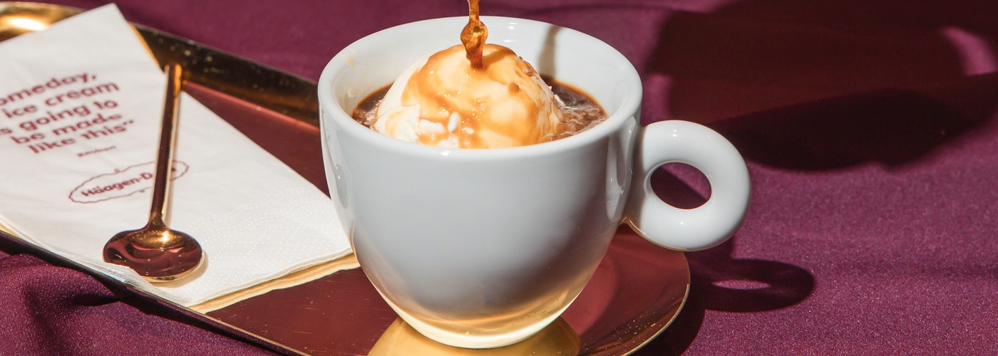 Fresh espresso being poured over a scoop of vanilla ice cream in a white cup, creating an affogato, served on a gold tray with a napkin and spoon on a deep burgundy surface.