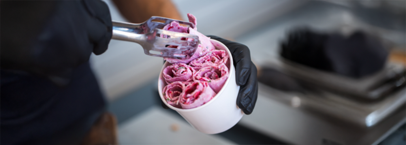 Close-up of a person wearing black gloves holding a white cup filled with pink rolled ice cream, using a metal utensil to arrange the rolls, with a blurred kitchen countertop in the background.