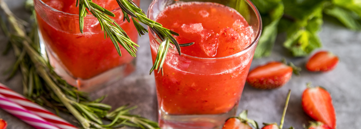 Two glasses of bright red strawberry mocktail with crushed ice, garnished with fresh rosemary sprigs, surrounded by sliced strawberries and herbs on a light surface.