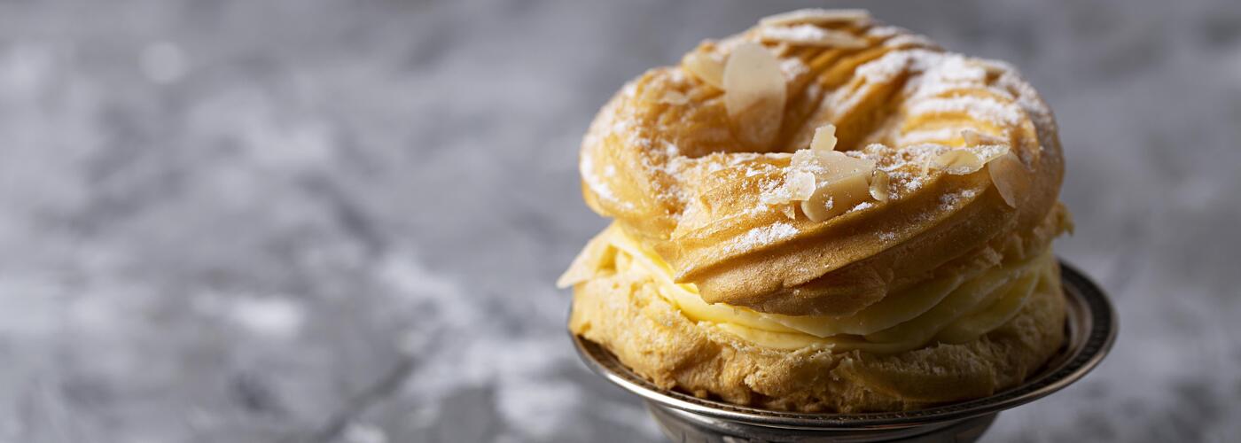 A golden, cream-filled choux pastry dusted with powdered sugar and almond flakes, placed on a small metal dish against a soft gray background.