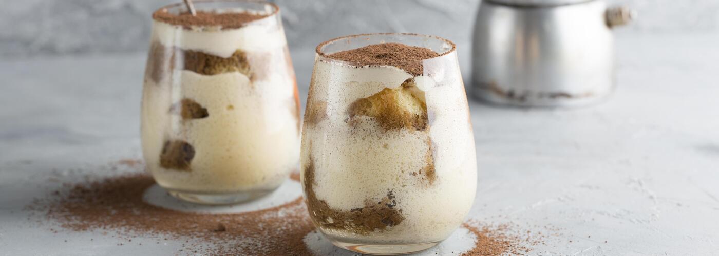 Two glasses of creamy tiramisu layered with mascarpone and soaked cake pieces, topped with a dusting of cocoa powder, placed on a light gray surface with cocoa scattered around.