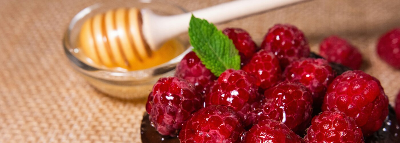Close-up of fresh raspberries topped with a mint leaf, with a honey dipper resting in a small glass bowl of honey in the background on a textured surface.